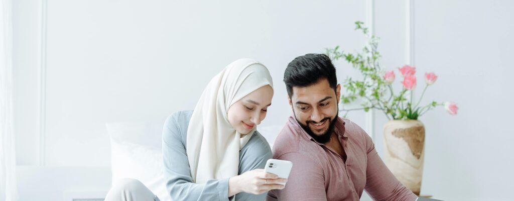 Young Muslim couple sitting back to back, using devices in a bright room.