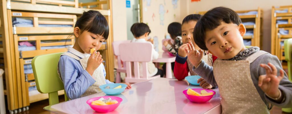 Kids seated around a table in a colorful classroom, eating snacks happily.
