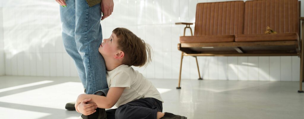 A young boy clings to a parent's leg indoors, expressing emotion in natural light.