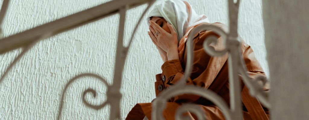 A woman in a hijab covers her face, depicting emotion on indoor stairs.