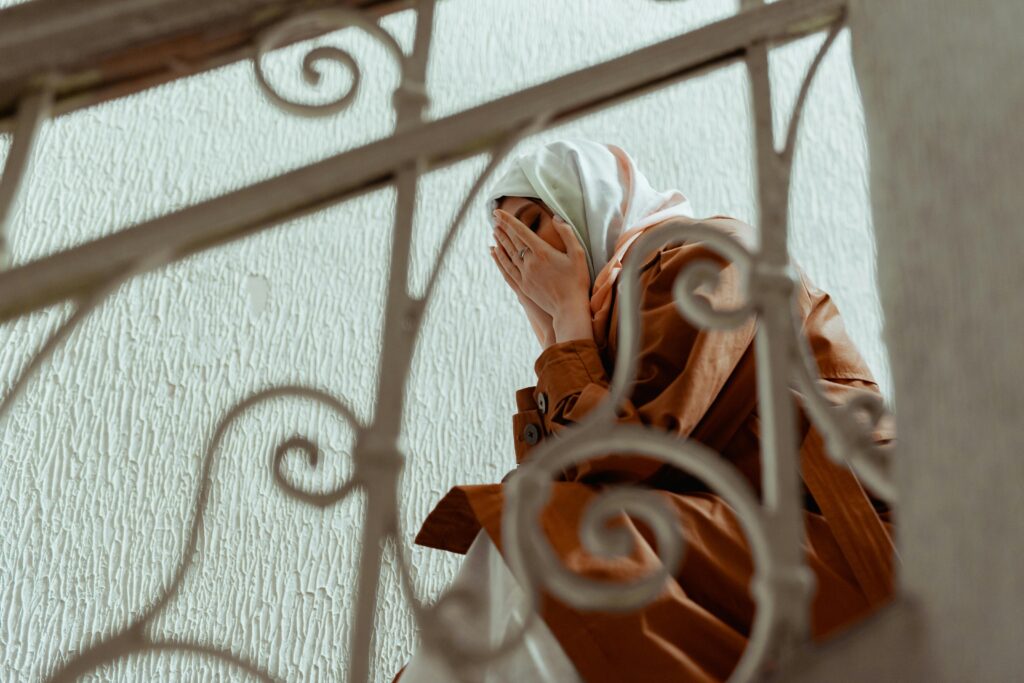A woman in a hijab covers her face, depicting emotion on indoor stairs.